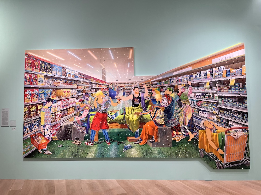 松山智一の大規模壁画作品。スーパーマーケットの店内風景を描いたポップアート。カラフルな衣装を着た多様な人物がショッピングカーを押したり、商品を手にしたりしている。背景には商品棚と陳列された食品パッケージが描かれており、現代社会の消費文化を表現している。白い壁に映える極彩色の作品