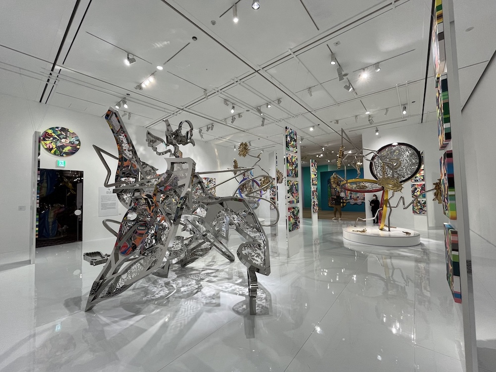 松山智一の抽象的な立体スカルプチャー。複雑に絡み合う金属製のパイプと幾何学的形態が3次元空間に展開している。ギャラリーの白い床と天井のある広い空間に設置された大規模インスタレーション。背景には壁に取り付けられた円形の彫刻作品も見える 